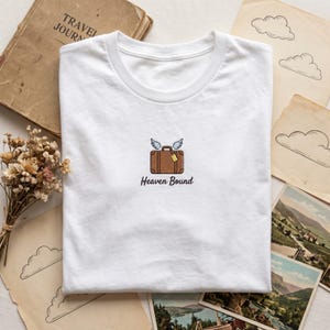 May include: A white t-shirt featuring a small embroidered graphic of a brown suitcase with light blue wings and a yellow luggage tag. Below the suitcase, the text "Heaven Bound" is embroidered in a black, cursive, lowercase font.