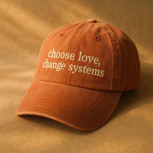May include: A burnt orange baseball cap featuring the embroidered text "choose love, change systems" in a light cream color. The cap has a structured crown, a curved brim with visible stitching, and small ventilation eyelets on the top panels.