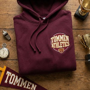 May include: A maroon hoodie with a drawstring hood and a circular patch that reads "TOMMEN ATHLETICS" in gold lettering. A stopwatch, whistle, and pennant are on a wooden surface.