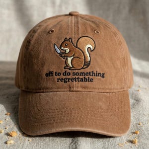 May include: A brown baseball cap with an embroidered design. The design features a squirrel holding a knife, with the text "off to do something regrettable" underneath. The squirrel is brown and white.