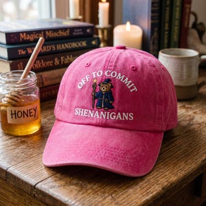 May include: A pink baseball cap with the text "OFF TO COMMIT SHENANIGANS" embroidered in white. The cap features a bear wearing a wizard's robe and hat. A jar of honey and books are in the background.