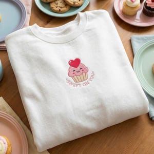 May include: White sweatshirt featuring an embroidered cupcake design. The cupcake is pink with a red heart and a smiling face. The words "Sweet on You" are embroidered around the cupcake in pink.