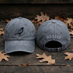 May include: Grey baseball cap featuring an embroidered crow on the front. The back of the cap has the word "RESIST" embroidered in white. The cap is displayed on a wooden surface with autumnal leaves.