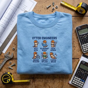 May include: A light blue crewneck sweatshirt featuring an embroidered graphic titled OTTER ENGINEERS. The design displays six cartoon otters wearing orange hard hats, labelled as BOLT BOSS, BLUEPRINT BUDDY, GEAR GENIUS, BRIDGE BUILDER, CIRCUIT SPLASH, and COFFEE BREAK.