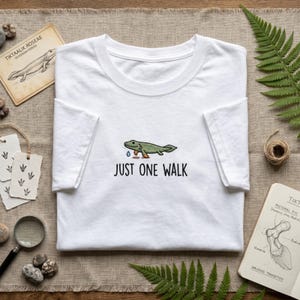May include: A white short-sleeve t-shirt featuring a graphic of a green fish-like creature with legs and the text "JUST ONE WALK" printed in black. The design includes a small blue water droplet icon positioned below the creature's head.