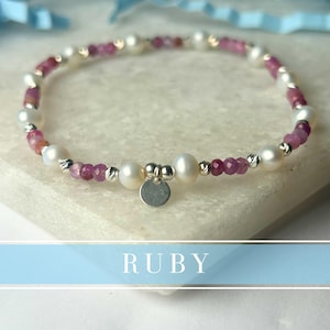 May include: A beaded bracelet featuring alternating faceted pinkish-red ruby gemstones and smooth white pearls. The bracelet includes small metallic spacer beads and a central circular silver-toned charm. The word RUBY is printed in white text.