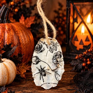 May include: An oval-shaped oyster shell ornament featuring a black ink illustration of roses and a spider. The shell has a rough, natural texture and is suspended by a piece of tan jute twine looped through a hole at the top of the shell.
