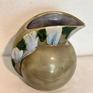 Puede incluir: Una pequeña jarra de cerámica con un esmalte verde, blanco y azul. La jarra tiene forma de concha y un cuerpo redondo.