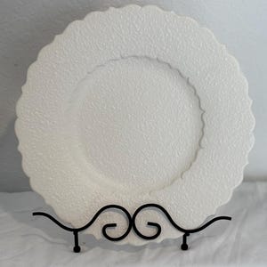 Könnte beinhalten: Weiße Keramik-Servierplatte mit strukturierter Oberfläche und gewelltem Rand. Die Platte steht auf einem schwarzen Metallständer.