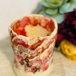 May include: A small, white ceramic planter with a unique, abstract design featuring red, brown, and yellow paint splatters. The planter has a slightly flared rim and a rounded base.