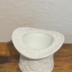 Puede incluir: Un recipiente blanco en forma de sombrero de copa de estilo vintage. El sombrero presenta un patrón texturizado detallado alrededor del borde y la base. El interior es liso y blanco. El recipiente está sobre una superficie de madera.