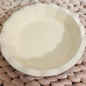 Op de afbeelding: Een crèmekleurige taartvorm met een geschulpte rand. De keramische schaal heeft een glad, glanzend oppervlak en een iets verhoogde rand. De taartplaat is rond en lijkt ongebruikt.