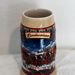 Può includere: Una boccale di birra Budweiser in ceramica con una scena natalizia che presenta una carrozza trainata da cavalli e un villaggio innevato. La boccale è decorata con colori rosso, verde e blu.