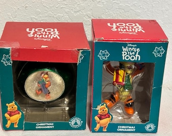 May include: Two Winnie the Pooh Christmas ornament boxes. The box on the left features a green ornament with a snow globe design of Tigger holding a gift. The box on the right features a clear ornament with Tigger holding a gift. Both boxes have a red background with the text "Disney's Winnie the Pooh" and "Christmas Ornament".