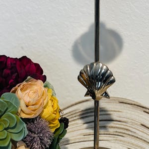 May include: Silver candlestick with a seashell design. The candlestick has a round base and a tall, slender stem. The seashell is located in the middle of the stem.