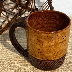 Puede incluir: Una taza de cerámica marrón con un esmalte moteado y un asa marrón oscuro. La taza tiene una banda de rayas marrones oscuras alrededor de la parte inferior.