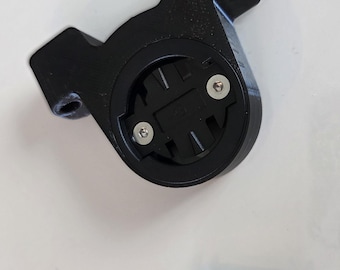 May include: Black plastic bicycle computer mount with two silver screws. The mount has a circular center with two screw holes and two arms extending outwards. The mount is designed to hold a bicycle computer.