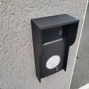 Può includere: Un campanello nero con un obiettivo della telecamera, un pulsante bianco con un'icona a forma di campana e due pulsanti più piccoli. Il campanello è montato su una parete grigia.