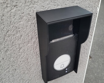 May include: A black doorbell with a camera lens, a white button with a bell icon, and two smaller buttons. The doorbell is mounted on a gray wall.