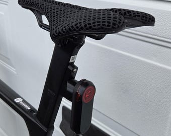 May include: A black bicycle saddle with a 3D-printed lattice structure mounted on a black seat post. A small, rectangular black rear light with a circular red lens is attached to the seat post below the saddle. The seat post features a QR code.
