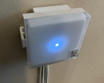 May include: A white square-shaped device with a blue light in the center. The device has a QR code on the side and is mounted on a wall.