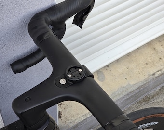 May include: Close-up of a black bicycle handlebar with black tape and a mounted black device. The handlebar curves upward and has a sleek, modern design. The device appears to be a mount for a cycling computer or other accessory.