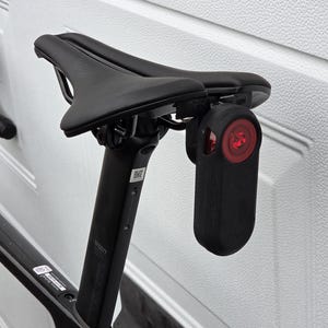 Op de afbeelding: Een zwart zadel van een fiets met een zwart achterlicht bevestigd aan de zadelpen. Het licht heeft een rode lens en een zwarte behuizing.