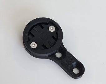 May include: A black bicycle computer mount with a circular top and an extended arm. The top has a recessed area with two silver screws and a central design. The arm has two holes for mounting.
