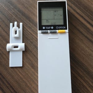 Może przedstawiać: Biały pilot do klimatyzacji z czarnym wyświetlaczem pokazującym czas, temperaturę i prędkość wentylatora. Pilot ma przyciski do regulacji temperatury, prędkości wentylatora i włączania/wyłączania. Obok pilota znajduje się biały plastikowy uchwyt ścienny.