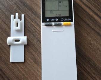 May include: White air conditioner remote control with a black display showing the time, temperature, and fan speed. The remote has buttons for temperature, fan speed, and on/off. There is a white plastic wall mount bracket next to the remote.