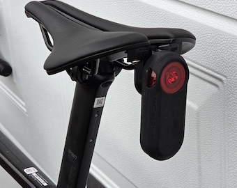 May include: A black bicycle seat with a black seat post and a red taillight attached. The seat has a perforated surface. The taillight is black and has a red light.