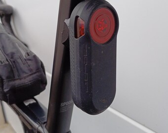 May include: A black bicycle tail light with a red lens is mounted on the seat post of a black bicycle. The light is rectangular with rounded edges. The bicycle frame is also black.