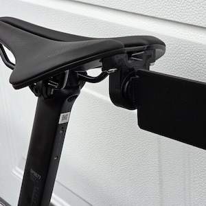 Peut inclure: Selle de vélo noire avec une fixation rectangulaire noire. La selle est montée sur une tige de selle noire. La selle a une surface texturée et un design épuré. La fixation semble être un support de caméra.