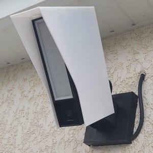 Peut inclure: Une caméra de sécurité extérieure noire avec un couvercle de protection blanc est montée sur un mur beige texturé. La caméra est de forme rectangulaire et la marque "Netatmo" est visible.