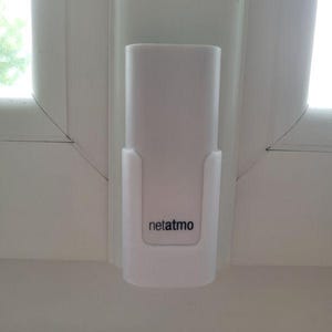 Peut inclure: Un appareil Netatmo blanc monté sur un cadre de fenêtre blanc. L'appareil est rectangulaire avec des bords arrondis et une petite zone encastrée en bas avec le nom de la marque "netatmo" imprimé en foncé.