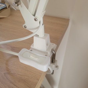 May include: White desk lamp with a clamp base and an inline switch. The lamp is attached to a wooden desk.