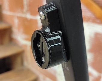 May include: Black plastic bicycle accessory, possibly a mount or holder, attached to a black frame. The accessory has a circular opening and is secured with a screw. A QR code sticker is visible on the frame.