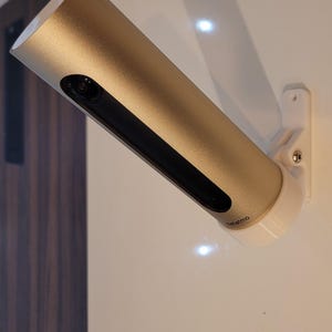 Peut inclure: Un appareil domotique cylindrique de couleur or, monté sur un mur blanc. L'appareil possède une zone de capteur rectangulaire noire et une base blanche. La marque "Netatmo" est visible.