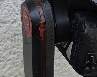 May include: Black and red Garmin bike light attached to a black bike helmet. The light has a rectangular shape with a red lens and a black body. The helmet also has a black saddle bag attached.