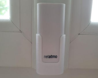 May include: A white Netatmo device mounted on a white window frame. The device is rectangular with rounded edges and a small, recessed area at the bottom with the brand name. The device is designed to monitor environmental conditions.