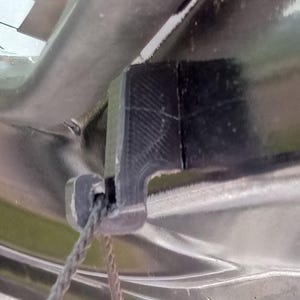 May include: Close-up of a black plastic component with a textured surface and a rope attached. The component appears to be part of a larger structure, possibly a vehicle or similar item. The rope is grey and appears to be made of woven fibres.