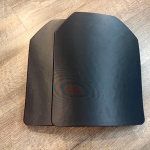 May include: Two black, rectangular ballistic plates with rounded corners are displayed. The plates have a slightly reflective surface and are positioned upright, side-by-side. The plates are designed to provide protection.