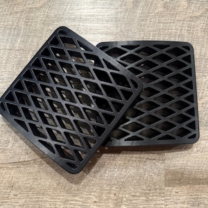 May include: Two black square-shaped mats with a diamond-shaped grid pattern. The mats are made of a durable material and are designed to provide traction or support. They are placed on a light brown wooden surface.