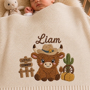 May include: A cream-colored knit baby blanket featuring an embroidered design of a brown highland cow wearing a cowboy hat, a green cactus, a hay bale, and a wooden sign that reads Little Cowboy, with the name Liam embroidered above the cow.