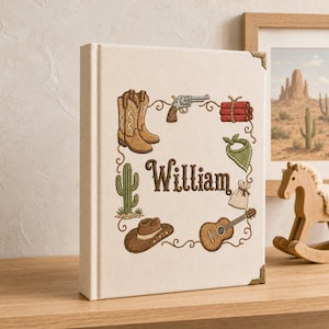 Op de afbeelding: Een crèmekleurig, met stof bekleed boek met metalen hoekbeschermers in goudkleur. De kaft is voorzien van geborduurde western-iconen, waaronder cowboylaarzen, een revolver, dynamiet, een cactus, een bandana, een zakje en een gitaar, met de naam William.