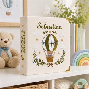 May include: A cream-colored photo album featuring embroidered details including the name Sebastian, the year 2026, a hot air balloon with a teddy bear in the basket, clouds, stars, and green leafy vines with small blue berries on the front cover.