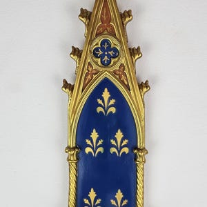 Puede incluir: Un estante de pared de color dorado con un diseño azul y dorado. El estante tiene un arco apuntado con una parte superior decorativa y un pequeño estante en la parte inferior.