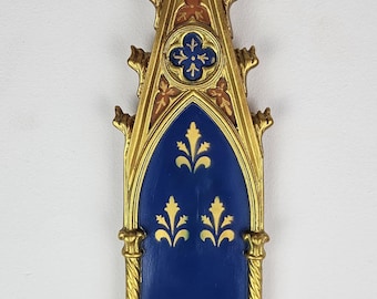 May include: A gold-colored wall shelf with a blue and gold design. The shelf has a pointed archway with a decorative top and a small shelf at the bottom.