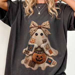 Può includere: Una t-shirt grigio antracite con una grafica centrale raffigurante un fantasma composto da vari quadrati di tessuto patchwork. Il fantasma ha occhi a bottone neri, un grande fiocco fantasia sulla testa e tiene in mano un piccolo secchiello a forma di zucca arancione.