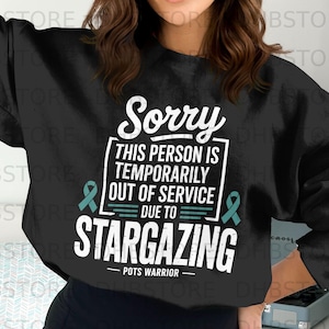 Puede incluir: Sudadera negra con texto blanco: "Sorry, THIS PERSON IS TEMPORARILY OUT OF SERVICE DUE TO STARGAZING." Cintas turquesas a ambos lados del texto. La parte inferior de la camiseta dice "POTS WARRIOR."